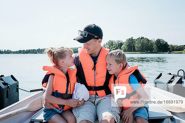Vater sitzt mit seinen Kindern auf einem Ruderboot im Sommer in Schweden und umarmt sie