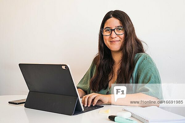 Junge spanische Brünette mit Brille  die mit ihrem Hybrid-Tablet-Laptop telearbeitet  um ihr Unternehmen an die neue Normalität nach der weltweiten Pandemie Covid 19 anzupassen. Sie schaut in die Kamera und lächelt