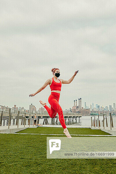Young female athlete jumping mid air wearing face mask by waterfront.