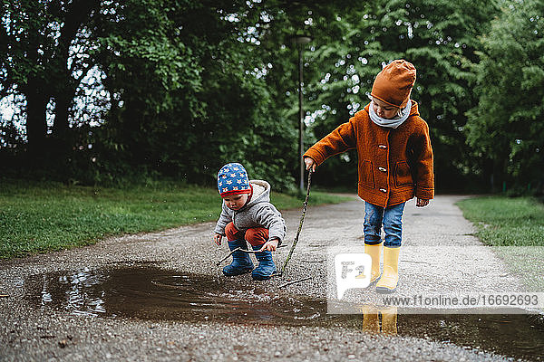Front view of young male kids playing with sticks in a puddle in park