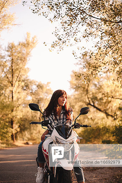 Confident young woman sitting on motorcycle on country road in forest