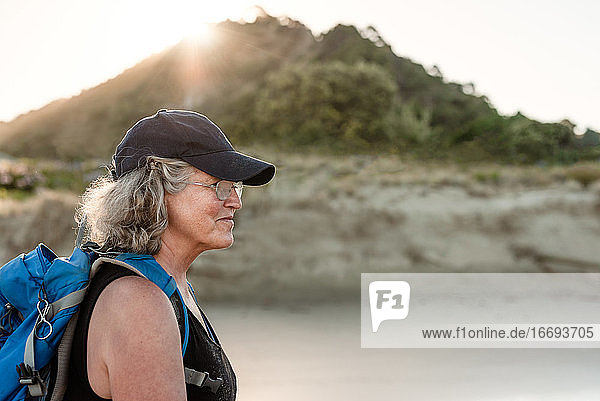 Aktive ältere Frau mit Rucksack und Hut  die an einem Strand in Neuseeland spazieren geht