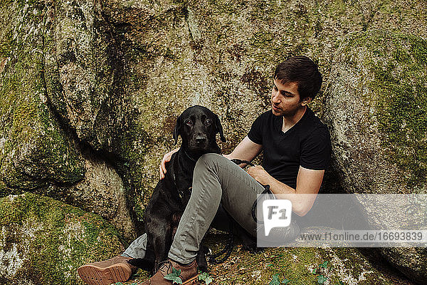 Junger Mann mit schwarzem Labrador Retriever auf einem moosbewachsenen Felsen sitzend