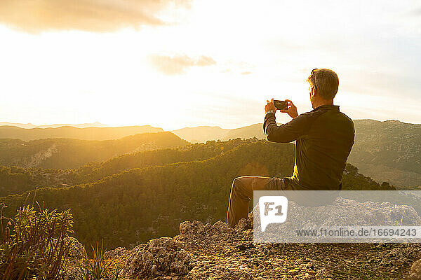 Man photographing the sunset with his mobile phone sitting on a rock