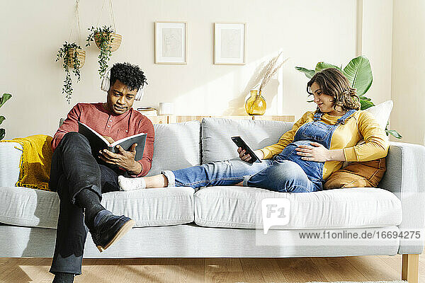 Husband listening to music and reading a book with his pregnant wife who is watching a tablet on a sofa. Interracial couple concept