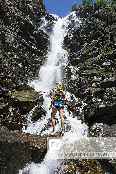 Rückansicht einer auf einem Felsen am Wasserfall stehenden Frau