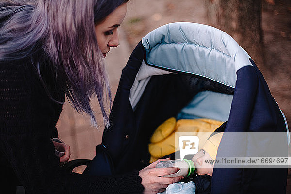 Mutter füttert ihren kleinen Sohn aus der Flasche im Kinderwagen im Park im Herbst