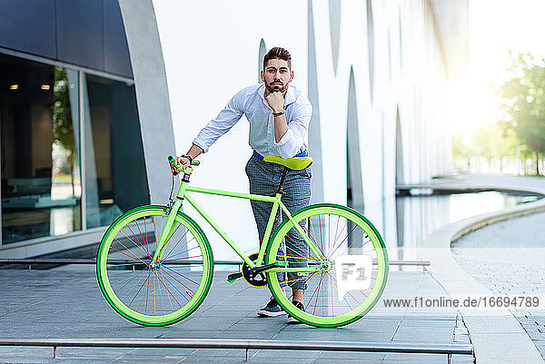Young bearded man leaning on bicycle seat while looking at camera