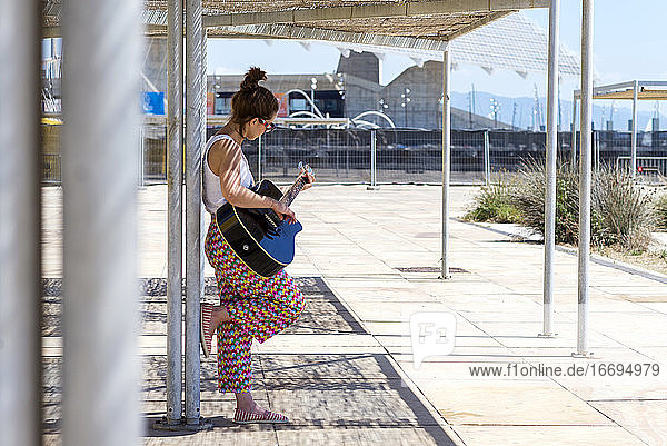 Junge Frau lehnt sich an eine Stange und spielt Gitarre im Freien