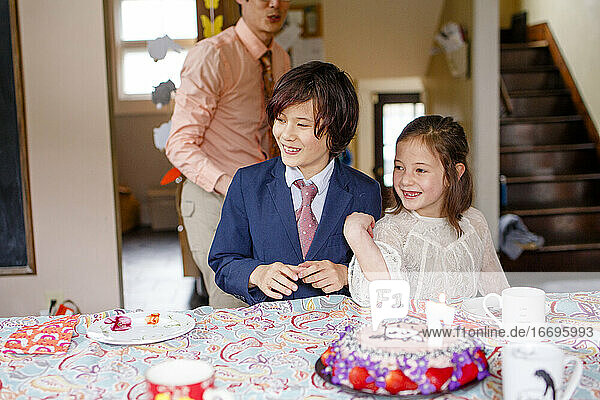 Smiling siblings sit at table in fancy clothes with lit birthday cake