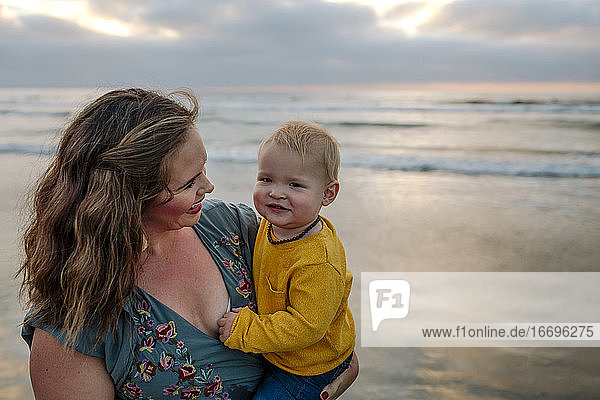 Schöne junge Mutter hält lächelnd 6 Monate altes Baby am Meer bei Sonnenuntergang