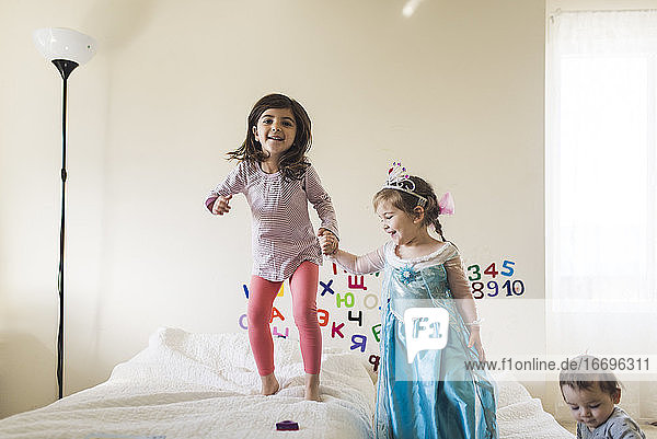 Lachendes 6-jähriges Mädchen und 4-jähriges Mädchen im Prinzessinnenkleid springen auf dem Bett
