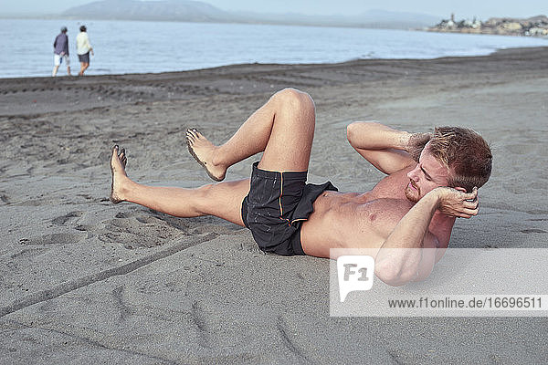 Hemdloser rothaariger junger Mann übt Bauch-Liegestütze am Strand