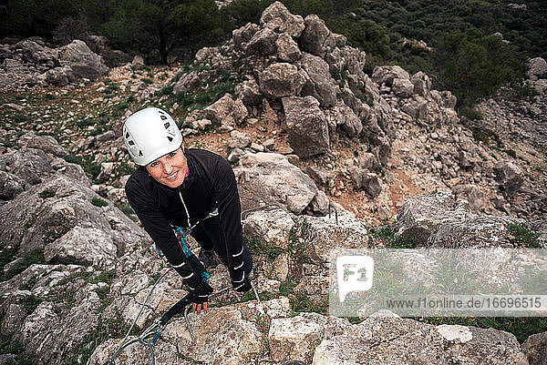 Concept: adventure. Climber woman with helmet and harness. Looking at camera happy. Anchored to the high natural wall. Smiling. Via ferrata in the mountains.