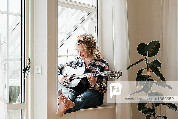Frau saß zu Hause auf einem Fenstersims und spielte lächelnd Gitarre