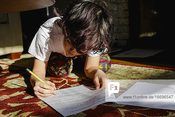 Ein Junge liegt auf einem Teppich in einem hellen Lichtfleck und macht Hausaufgaben
