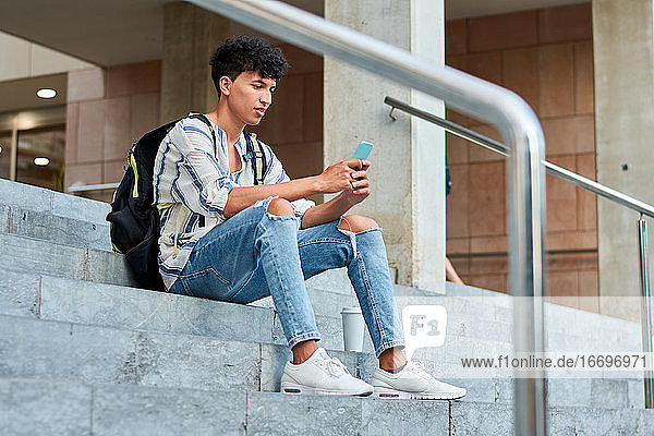 Young afro uses his mobile while he is sitting on the stairs