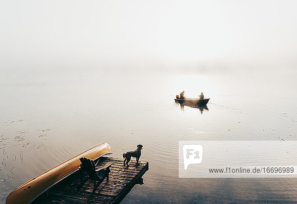 Zwei Personen in einem Fischerboot im Nebel mit einem Hund  der vom Steg aus zusieht.