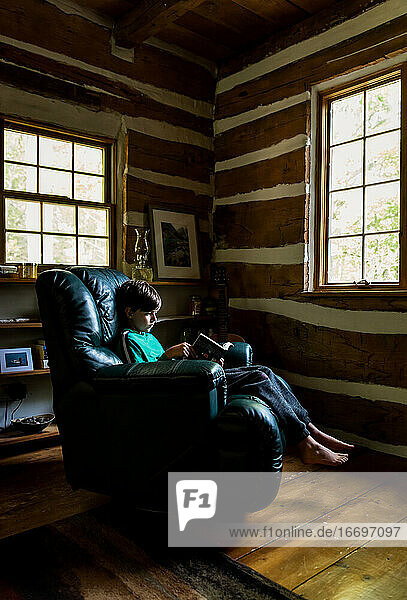 Kleiner Junge liest im Ledersessel in einem rustikalen Blockhaus.