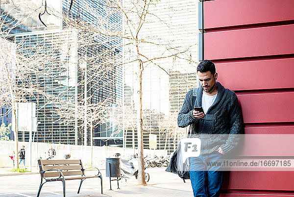 Vorderansicht eines bärtigen Mannes  der ein Telefon benutzt und sich an die Wand eines Bürogebäudes lehnt