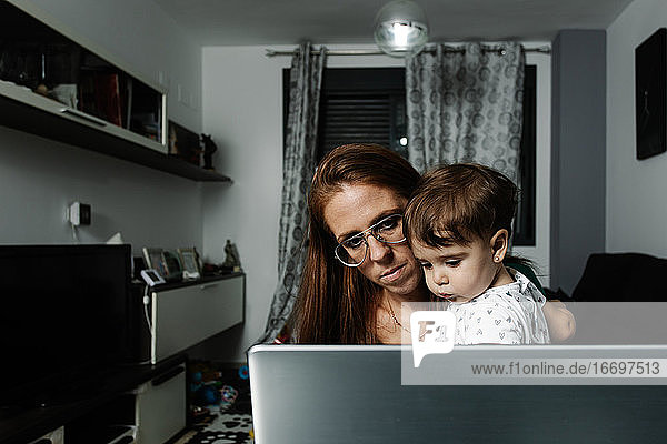 Frau mit kleinem Kind auf dem Arm benutzt Laptop zu Hause