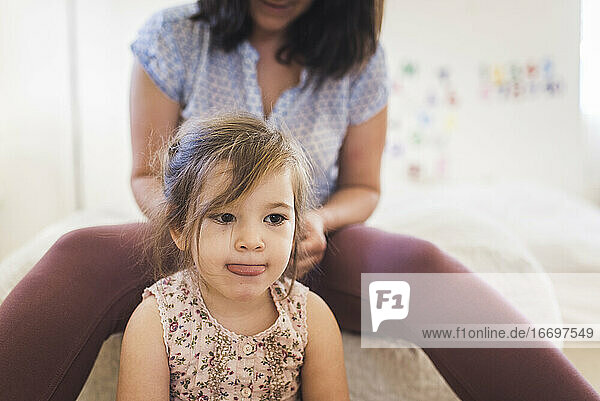 Mom sitting on bed braiding hair of 4 yr old daughter with tongue out