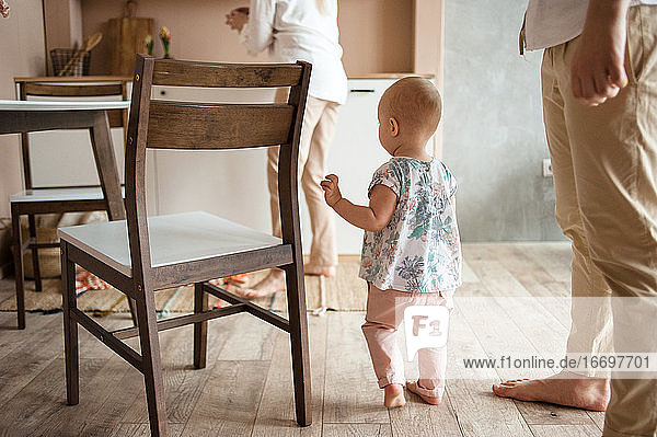 Kleines Baby geht durch Holzboden in der Küche zwischen den Beinen der Eltern.