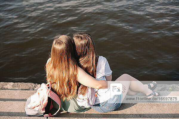 Rückenansicht eines lesbischen Paares  das sich umarmt  während es auf Stufen am Fluss sitzt