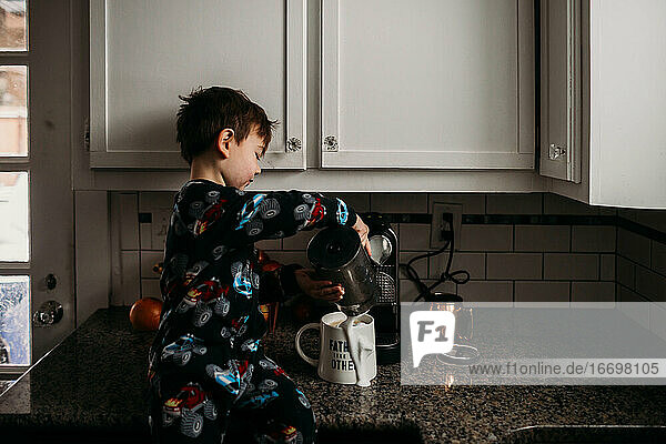 Junge sitzt auf Küchenzeile und verschüttet Milch auf Kaffeetasse