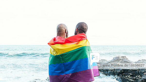 Lgbt Stolz Konzept. Homosexuelles Paar  das sich liebevoll umarmt  eingewickelt in die Regenbogenflagge mit Blick auf die Meereslandschaft.