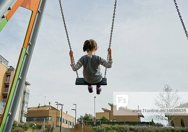 back view of happy little girl swinging high on playground swing set