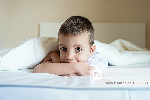 Adorable kid lying down on bed under blanket  while looking camera