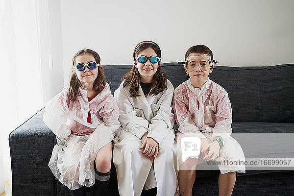 Fröhliche Kinder mit Schutzbrillen und Schutzmänteln sitzen auf dem Sofa