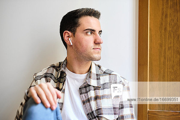 Porträt eines jungen Mannes  der zu Hause chillt und wegschaut  während er mit Kopfhörern Musik hört.