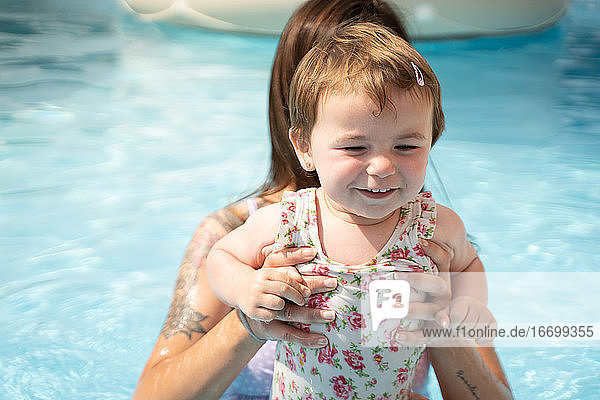 Kleines Mädchen lächelnd mit den Händen einer Frau in der Mitte eines Pools