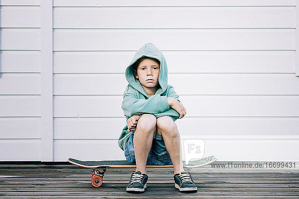 young boy sat on a skateboard with a hoodie on looking grumpy