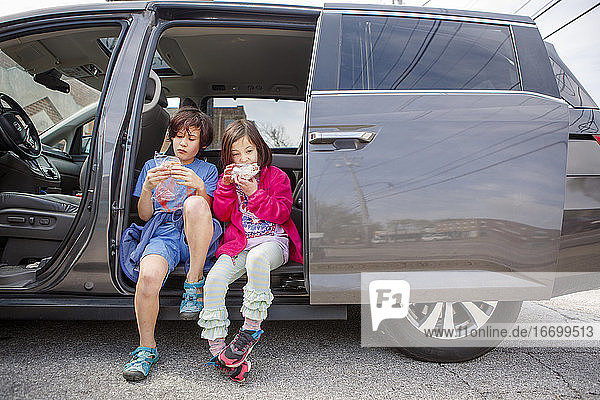 Two children sit side-by-side in an open van eating together