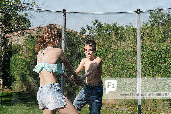 Horizontales Foto eines blonden Jungen und eines brünetten Mädchens  die sich an den Händen halten und auf einem Trampolin auf einem grünen Spielplatz spielen. Der Teenager und das Kind tragen kurze Jeans und sie sehen glücklich aus  lächelnd