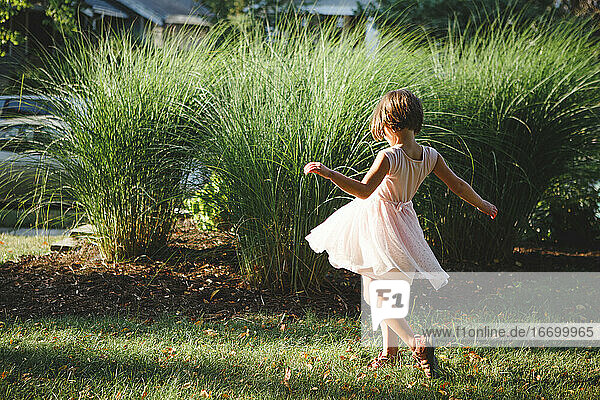 A small child in a soft pink dress twirls in front of long green grass