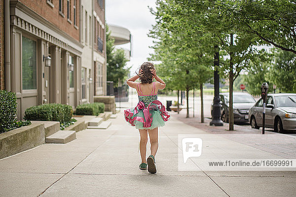 Ein kleines Mädchen läuft im Tütü auf dem Bürgersteig und hält sein Haar gegen den Wind