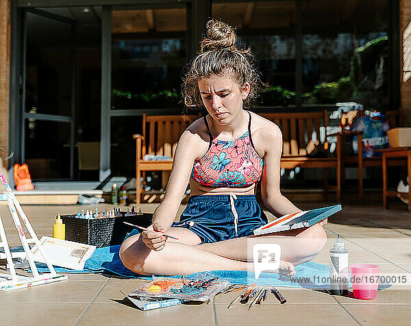 weißes Mädchen malen ein Bild sitzt auf einer Terrasse mit allen Malerei Elemente um wie Pinsel  Farben  Wasser .