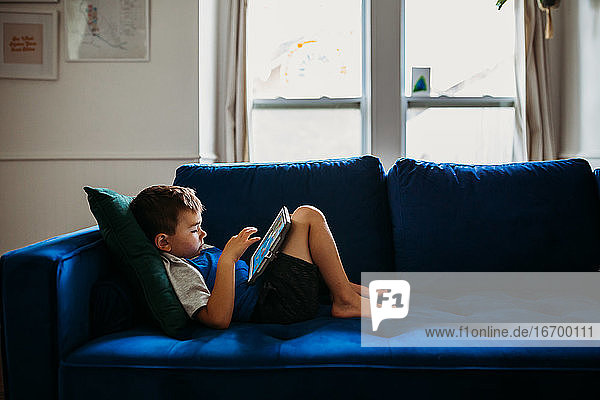 Young boy laying on blue couch playing educational games on tablet