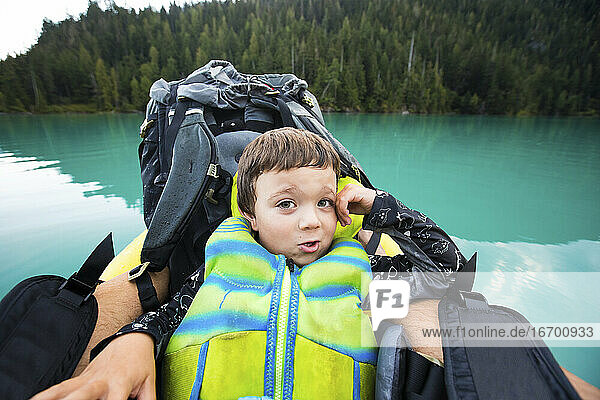 Boy wearing PFD rests against backpack on inflatable boat on blue lake