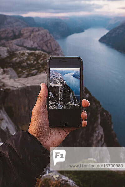 Hand hält Smartphone mit Bild der Szene im Hintergrund auf sie an norwegischen Fjorden.
