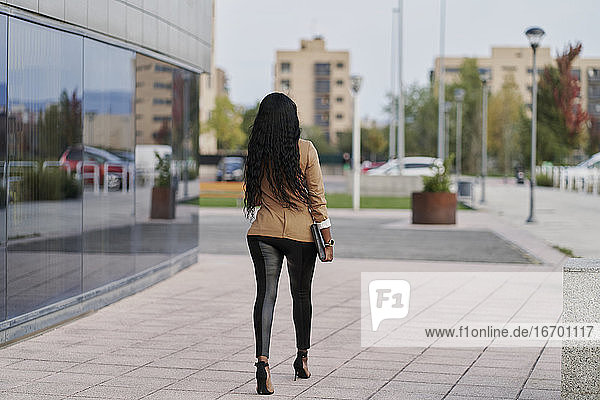 Rückenansicht einer schwarzen Frau in einem braunen Anzug  die einen Laptop und einen Behälter mit Kaffee in der Hand hält und neben einem Gebäude spazieren geht. Geschäftskonzept