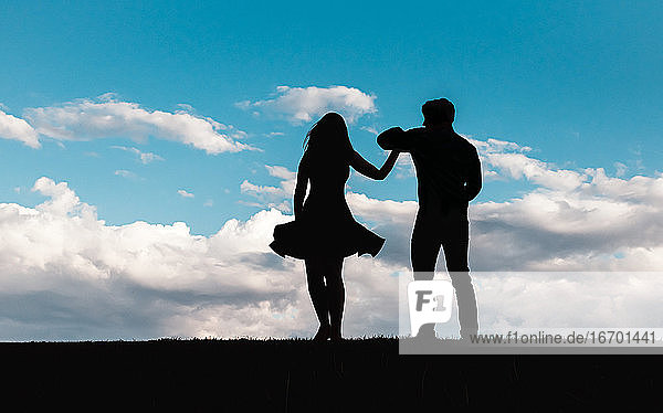 Silhouette of man and woman dancing against a blue sky with clouds.