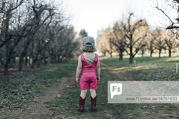 Ein junges Mädchen steht in einem Obstgarten und trägt ein Trikot und Cowgirl-Stiefel.
