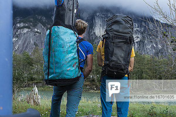 Zwei Menschen mit großem Rucksack stehen vor einem Berg und einem Fluss