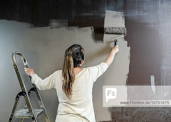 Frau klettert auf eine Leiter und fängt eine Farbrolle voller grauer Farbe auf einer braunen Wand. Der Maler fährt die Rolle hoch und runter und bedeckt die Wand mit grauer Farbe  die noch feucht ist. Horizonta