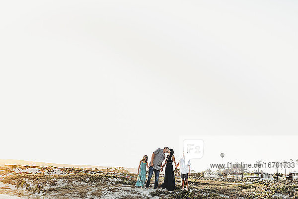 Familie hält sich am Strand an den Händen  während der Vater die Mutter küsst